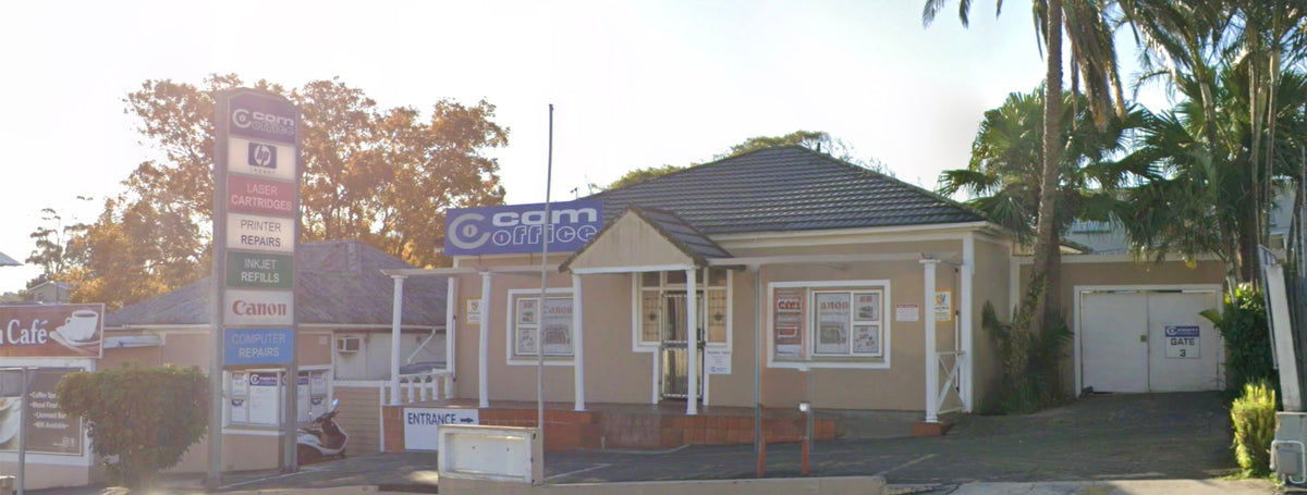 A small beige single-story commercial building with a dark tiled roof, white trim, and a blue sign reading 'ccm office', shown outdoors with trees, a palm tree, and a tall retail signpost at left.
